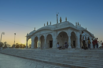 A white marble temple stands majestically with intricate architectural details, adorned with multiple domes and decorative spires. A group of people is gathered around the steps, some sitting and others standing, while the sun sets in the background, casting a warm glow on the scene.