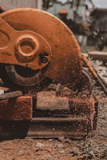 A close-up view of an industrial circular saw in motion, cutting through metal or a similar material, producing sparks that fly in various directions. The saw is predominantly orange and appears to be weathered. The surrounding area has a gritty, construction-like environment with scattered debris.