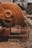 A close-up view of an industrial circular saw in motion, cutting through metal or a similar material, producing sparks that fly in various directions. The saw is predominantly orange and appears to be weathered. The surrounding area has a gritty, construction-like environment with scattered debris.