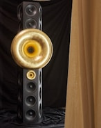 A tall, rectangular speaker tower with multiple circular speaker units. Two large, golden horn-like structures are mounted on the front, creating a striking visual contrast against the black speakers and drapery.
