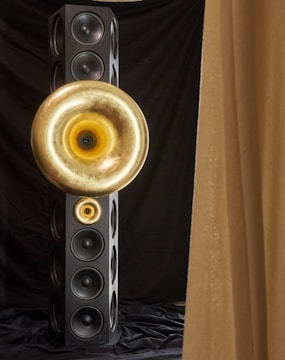 A tall, rectangular speaker tower with multiple circular speaker units. Two large, golden horn-like structures are mounted on the front, creating a striking visual contrast against the black speakers and drapery.