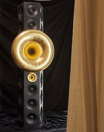 A tall, rectangular speaker tower with multiple circular speaker units. Two large, golden horn-like structures are mounted on the front, creating a striking visual contrast against the black speakers and drapery.