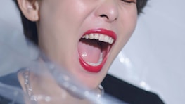 A close-up image of a person with their mouth open wide, showing red lipstick on the lips and white teeth. The person appears to be expressing a strong emotion, possibly yelling or singing. The background is soft and out of focus, with hints of light reflecting off surfaces.