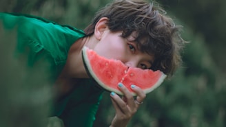 A person with short, curly hair is holding a slice of watermelon in front of their face, revealing only one eye. The background is lush and green, and the person is wearing a green garment. Their nails are painted a light blue color.