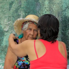 An elderly woman wearing a straw hat and colorful clothing is embraced by another person in a red tank top. The woman has an expression that appears contemplative or thoughtful. The background is textured in shades of green.