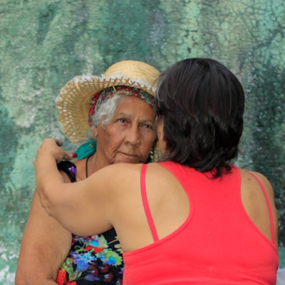An elderly woman wearing a straw hat and colorful clothing is embraced by another person in a red tank top. The woman has an expression that appears contemplative or thoughtful. The background is textured in shades of green.