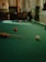 A person is holding a pool cue on a billiard table, with several colored balls visible. The setting is a dimly lit room with wooden and green details in the background.