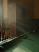 A dimly lit room with a pool table in partial focus. Sunlight streams through, creating a warm, inviting atmosphere. Some pool balls are positioned on the table. An old wooden cabinet and bar stools are faintly visible in the background.