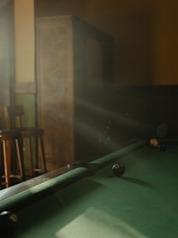 A dimly lit room with a pool table in partial focus. Sunlight streams through, creating a warm, inviting atmosphere. Some pool balls are positioned on the table. An old wooden cabinet and bar stools are faintly visible in the background.
