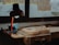 A wooden guitar body lies on a workbench near a window. A lamp with a red stem illuminates the area, casting a soft glow over the workspace. Various woodworking tools and materials are scattered around the bench. The window provides a view of an outdoor space with a blurred figure in the background.