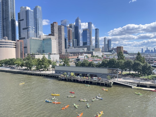 A group of kayakers paddling on a river in front of a city skyline filled with tall skyscrapers. The scene includes a dock with greenery nearby, and the sky is bright with scattered clouds.