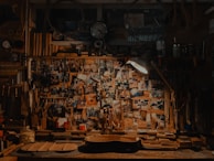 A cluttered workshop filled with woodworking tools, photos pinned on a bulletin board, and a guitar body in progress on the workbench. The scene is dimly lit by a lamp, adding a cozy, vintage feel to the space.