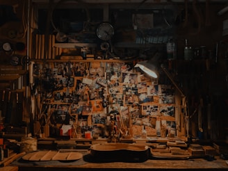A cluttered workshop filled with woodworking tools, photos pinned on a bulletin board, and a guitar body in progress on the workbench. The scene is dimly lit by a lamp, adding a cozy, vintage feel to the space.