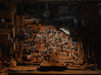 A cluttered workshop filled with woodworking tools, photos pinned on a bulletin board, and a guitar body in progress on the workbench. The scene is dimly lit by a lamp, adding a cozy, vintage feel to the space.