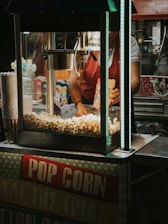A popcorn stand with a machine overflowing with freshly popped corn. A person, partially visible, is operating the machine and holding a popcorn bag. The surrounding area includes popcorn containers and colorful packaging.