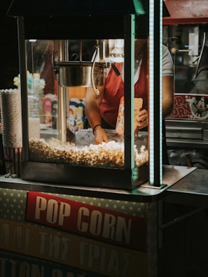 A popcorn stand with a machine overflowing with freshly popped corn. A person, partially visible, is operating the machine and holding a popcorn bag. The surrounding area includes popcorn containers and colorful packaging.