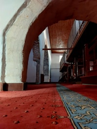 An interior space with traditional architecture featuring an arched stone structure and a richly patterned carpet covering the floor. The walls are made of a mix of stone and plaster, and there is a wooden ceiling supported by beams. A balcony with ornate railings runs along one side.