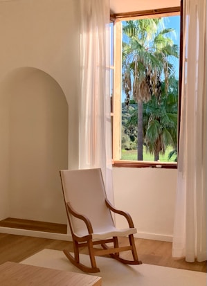 A serene room featuring a wooden rocking chair placed near an open window. The window has thin white curtains, allowing sunlight to brighten the space. Outside, lush palm trees can be seen against a clear blue sky, creating a calming natural view.