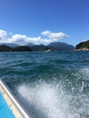 A vibrant seascape featuring clear blue waters with visible waves, surrounding green islands with hilly terrain, and a bright sky populated with fluffy white clouds. The foreground shows a portion of a boat, partially submerged in water and creating a splash.