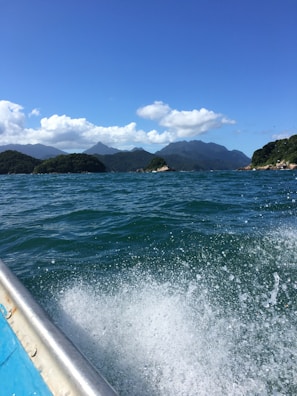 A vibrant seascape featuring clear blue waters with visible waves, surrounding green islands with hilly terrain, and a bright sky populated with fluffy white clouds. The foreground shows a portion of a boat, partially submerged in water and creating a splash.