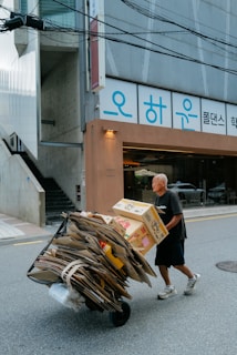 An elderly man is pushing a cart loaded with flattened cardboard boxes along a city street. The setting is urban, with a building featuring Korean signage in the background. The street is empty aside from the man, and the light suggests it is daytime.