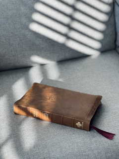 A brown leather-bound book rests on a gray fabric surface. Sunlight filters through blinds, casting striped shadows across the book. The book's spine reads 'Holy Bible,' and a red ribbon bookmark peeks out from the bottom.