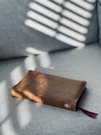 A brown leather-bound book rests on a gray fabric surface. Sunlight filters through blinds, casting striped shadows across the book. The book's spine reads 'Holy Bible,' and a red ribbon bookmark peeks out from the bottom.
