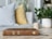 A brown Bible with the text 'Holy Bible: English Standard Version' embossed on the spine rests on a light-colored wooden surface. In the background, there is a striped pillow, a yellow cushion, a small potted green plant, and a cup. The setting appears calm and cozy, indicating a domestic or contemplative environment.