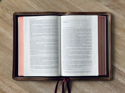 An open Bible lies on a wooden surface, displaying the books of 2 Chronicles and Ezra. The pages have a white background with black text, and the edges of the pages are stained red. A brown leather cover borders the Bible, and two maroon ribbons are visible.