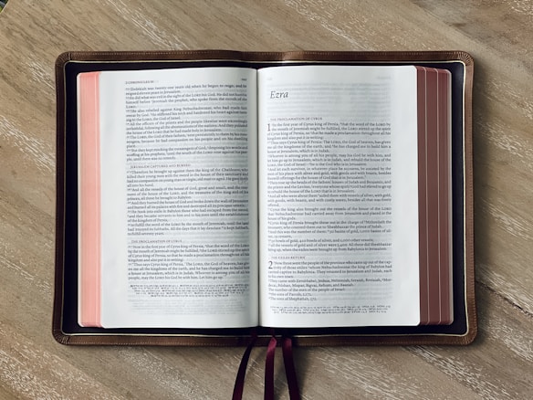 An open Bible lies on a wooden surface, displaying the books of 2 Chronicles and Ezra. The pages have a white background with black text, and the edges of the pages are stained red. A brown leather cover borders the Bible, and two maroon ribbons are visible.