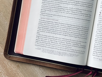 An open book is displayed with text in a serif font on its pages. The book appears to have religious or historical content, likely a Bible, as suggested by the chapter and verse notation. The pages have a pink ribbon and are set within a brown leather cover. The book lies on a wooden surface.
