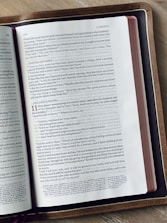 A close-up of an open book placed on a wooden surface. The book appears to be a Bible, featuring text primarily in black font on white pages with a reference to 'LUKE 10-11'. The edges of the book have a red tint, and the book has a brown leather cover.