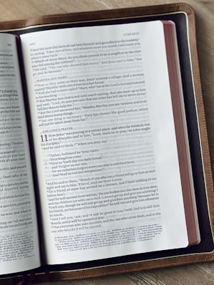 A close-up of an open book placed on a wooden surface. The book appears to be a Bible, featuring text primarily in black font on white pages with a reference to 'LUKE 10-11'. The edges of the book have a red tint, and the book has a brown leather cover.