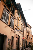 A narrow European street lined with yellow-toned buildings featuring green shuttered windows. A sign reading 'TRATTORIA' hangs from one of the buildings, while a small Italian flag is displayed prominently. Bikes are parked casually along the sidewalk, and potted plants add a touch of greenery.
