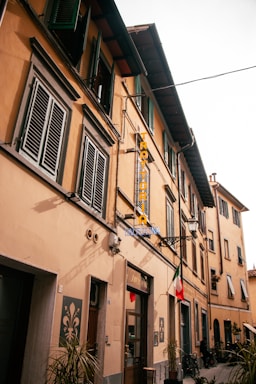 A narrow European street lined with yellow-toned buildings featuring green shuttered windows. A sign reading 'TRATTORIA' hangs from one of the buildings, while a small Italian flag is displayed prominently. Bikes are parked casually along the sidewalk, and potted plants add a touch of greenery.