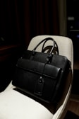 A black leather briefcase with a luggage tag is positioned on a light-colored chair. The background is dimly lit, providing a contrast with the dark bag, which has two handles and a front flap.