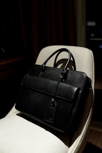 A black leather briefcase with a luggage tag is positioned on a light-colored chair. The background is dimly lit, providing a contrast with the dark bag, which has two handles and a front flap.
