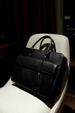 A black leather briefcase with a luggage tag is positioned on a light-colored chair. The background is dimly lit, providing a contrast with the dark bag, which has two handles and a front flap.