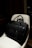 A black leather briefcase with a luggage tag is positioned on a light-colored chair. The background is dimly lit, providing a contrast with the dark bag, which has two handles and a front flap.