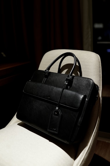 A black leather briefcase with a luggage tag is positioned on a light-colored chair. The background is dimly lit, providing a contrast with the dark bag, which has two handles and a front flap.