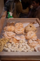 A display of various types of fresh pasta in a wooden frame, including fettuccine, fusilli, and gnocchi. The pasta is arranged in neat rows and piles, showcasing different shapes and textures. In the background, a spray bottle is visible, and there are blurred figures of people, suggesting an indoor market or shop setting.