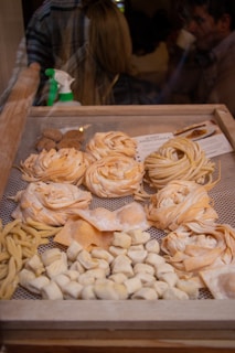 A display of various types of fresh pasta in a wooden frame, including fettuccine, fusilli, and gnocchi. The pasta is arranged in neat rows and piles, showcasing different shapes and textures. In the background, a spray bottle is visible, and there are blurred figures of people, suggesting an indoor market or shop setting.