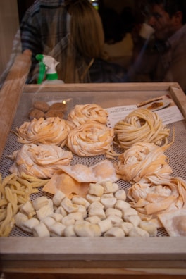 A display of various types of fresh pasta in a wooden frame, including fettuccine, fusilli, and gnocchi. The pasta is arranged in neat rows and piles, showcasing different shapes and textures. In the background, a spray bottle is visible, and there are blurred figures of people, suggesting an indoor market or shop setting.