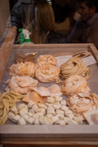 A display of various types of fresh pasta in a wooden frame, including fettuccine, fusilli, and gnocchi. The pasta is arranged in neat rows and piles, showcasing different shapes and textures. In the background, a spray bottle is visible, and there are blurred figures of people, suggesting an indoor market or shop setting.