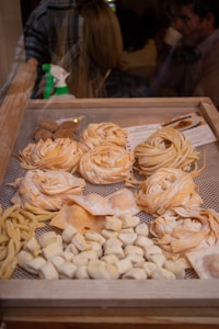 A display of various types of fresh pasta in a wooden frame, including fettuccine, fusilli, and gnocchi. The pasta is arranged in neat rows and piles, showcasing different shapes and textures. In the background, a spray bottle is visible, and there are blurred figures of people, suggesting an indoor market or shop setting.