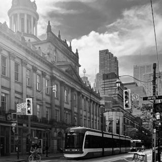 An urban street scene featuring a large, historic building with a grand architectural style. In the foreground, a modern streetcar travels along the road. People are walking and a person is riding a bicycle. Tall, contemporary skyscrapers rise in the background, under a partially cloudy sky.