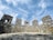 A stone wall stands prominently under a partly cloudy sky. The wall is constructed with rough, irregular stones and includes several rectangular openings resembling windows or battlements. The foreground is paved with similarly textured stones, creating a historical or ancient ambiance.