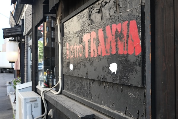 A rustic wooden exterior with a weathered sign displaying the words 'Bistro TRAMA' in bold red letters. The setting suggests a cozy, inviting atmosphere with visible bottles and an air conditioning unit nearby.