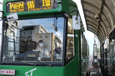 A green tram is stationed at a covered tram stop with a driver wearing a face mask visible through the front window. The tram displays 'Inner loop' and 'circular route' in both Japanese and English on an electronic sign. The surrounding environment shows residential buildings and clear skies.