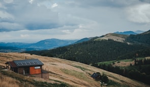 A small wooden cabin with solar panels is situated on a hillside, surrounded by rolling green hills and dense forests. The sky above is overcast with patches of clouds, creating a moody atmosphere. The scene is tranquil and isolated, with vast expanses of untouched nature in the background.